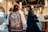 Three men are standing in a well-lit indoor space, possibly a market or showroom. One man is wearing a jacket with a map of Japan printed on the back, while the other two wear hats and casual attire. The setting includes large hanging lights and various booths and displays.