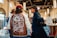 Three men are standing in a well-lit indoor space, possibly a market or showroom. One man is wearing a jacket with a map of Japan printed on the back, while the other two wear hats and casual attire. The setting includes large hanging lights and various booths and displays.