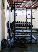 A gym setting with various equipment including a power rack, barbell, weight plates, and a row of dumbbells against a wall. The room has white brick walls and a wooden ceiling with visible beams. Natural light is entering through small windows.