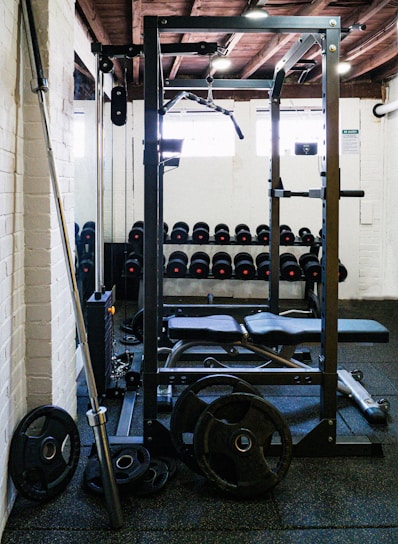 A gym setting with various equipment including a power rack, barbell, weight plates, and a row of dumbbells against a wall. The room has white brick walls and a wooden ceiling with visible beams. Natural light is entering through small windows.