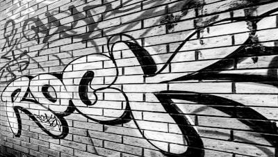 A brick wall features bold graffiti art with the word 'Rock' painted prominently in black and white. The wall is composed of rectangular bricks arranged in a horizontal pattern with some weathering and other graffiti tags visible in the background.