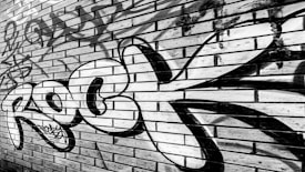 A brick wall features bold graffiti art with the word 'Rock' painted prominently in black and white. The wall is composed of rectangular bricks arranged in a horizontal pattern with some weathering and other graffiti tags visible in the background.