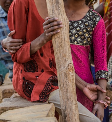 A group of people wearing colorful clothing, including a red garment and a patterned dress, standing close together. Their hands are visible, one of which is holding onto a wooden stick. The scene appears to be casual and outdoors.
