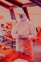 A person wearing full protective medical gear and a face shield is holding a clipboard and pen in a tent-like environment. Various equipment and items are scattered on tables in the background.