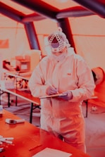 A person wearing full protective medical gear and a face shield is holding a clipboard and pen in a tent-like environment. Various equipment and items are scattered on tables in the background.