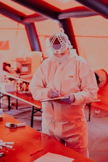 A person wearing full protective medical gear and a face shield is holding a clipboard and pen in a tent-like environment. Various equipment and items are scattered on tables in the background.