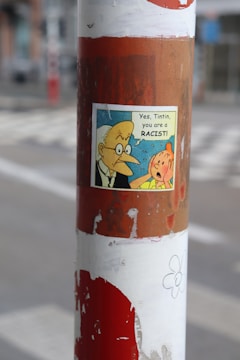 A street pole with a weathered, peeling sticker featuring a comic strip image. The comic panel includes two cartoon characters with speech bubbles, and the background displays faded urban scenery.