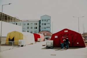 A series of colorful tents are set up outside a modern building with multiple windows. The tents are red, yellow, and cream, and are surrounded by barriers and equipment. Two people stand near a red tent, engaged in conversation. The setting appears to be an organized outdoor area, possibly for events or emergency situations.