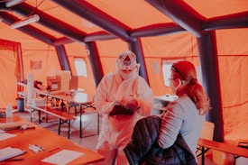 Inside an orange tent, a person in full personal protective equipment speaks with another masked individual. Various tables hold supplies and paper documents, creating an organized setup.