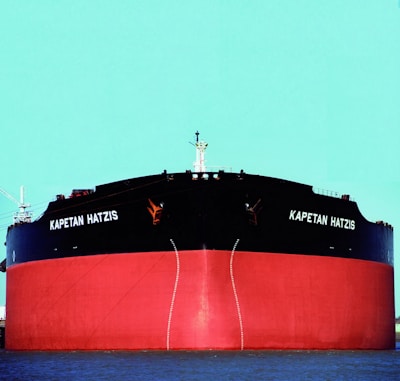 A large cargo ship with the name 'Kapetan Hatzis' is prominently displayed on the front. The ship's body is painted black with a red lower section. It is situated against a clear turquoise sky and is partially submerged in water.