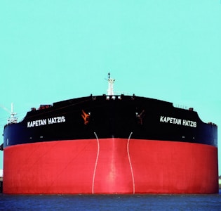 A large cargo ship with the name 'Kapetan Hatzis' is prominently displayed on the front. The ship's body is painted black with a red lower section. It is situated against a clear turquoise sky and is partially submerged in water.