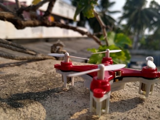 A small red and white quadcopter drone rests on a rough, concrete surface near some intertwined ropes and branches. In the background, blurred palm trees and foliage are visible, suggesting an outdoor setting.