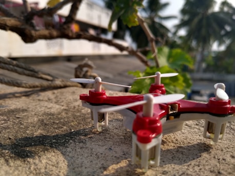 A small red and white quadcopter drone rests on a rough, concrete surface near some intertwined ropes and branches. In the background, blurred palm trees and foliage are visible, suggesting an outdoor setting.