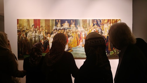 A group of people are observing a large, classical painting in a gallery setting. The painting depicts a historical or regal scene with numerous figures in elaborate clothing. The observers are silhouetted against the brightly lit artwork.