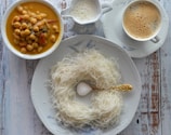 A breakfast setting with a plate of string hoppers and sugar on a decorative spoon, a bowl of chickpea curry, a cup of frothy coffee, and a milk jug all placed on a rustic wooden table.