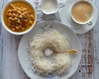 A breakfast setting with a plate of string hoppers and sugar on a decorative spoon, a bowl of chickpea curry, a cup of frothy coffee, and a milk jug all placed on a rustic wooden table.