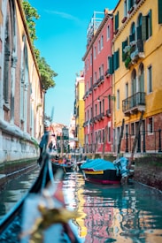 A picturesque canal flanked by colorful buildings with balconies and shuttered windows. Gondolas float gently on the clear water, reflecting the vibrant facades. The scene is bathed in bright daylight, with a vivid blue sky above.