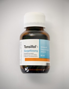 A brown glass bottle with a white cap is labeled with the name 'Tonsillol' and details about the contents, indicating it is a gargling solution against inflammation and infections in the mouth and throat area. The label includes details like the active ingredient Dequalinium chloride and the volume of 60 ml.