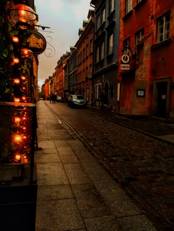 A dimly lit cobblestone street flanked by old European-style buildings with colorful facades. Warm, glowing lanterns line one side of the street, creating a cozy ambiance. A few pedestrians and a parked car are visible in the distance.