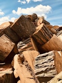 A large stack of freshly cut logs with visible bark and rough textures against a backdrop of a blue sky scattered with white clouds.