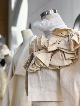 A close-up view of a dress form draped with a beige fabric, featuring detailed ruffles and pleats around the neckline. The fabric appears to be a natural or linen material, with visible texture. Soft lighting enhances the fabric's texture, creating subtle shadows.