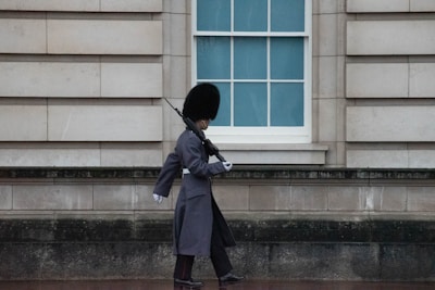 A guard in a traditional uniform and bearskin hat marches beside a building with large stone blocks. The guard carries a rifle over the shoulder and walks in front of a window with a blue tint.