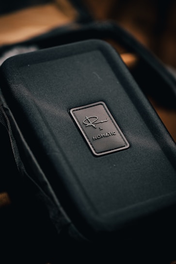 A close-up of a textured, dark-colored rectangular case, featuring a prominent logo with the text 'NOMATIC' on a small badge in the center.