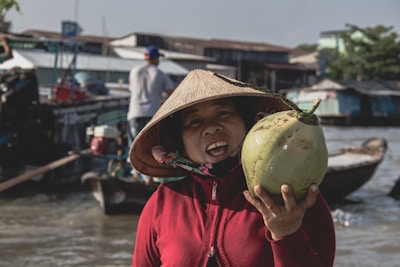 A person wearing a conical hat is holding a large green coconut and smiling. They are in an outdoor market setting with boats and water in the background. The surrounding area includes various structures and other people engaged in daily activities.
