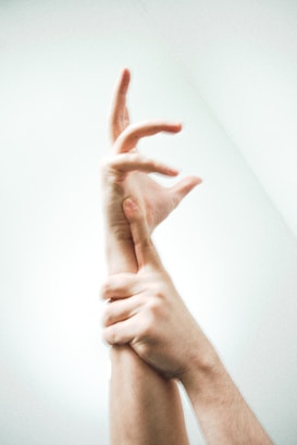 A pair of hands, one grasping the wrist of the other, with fingers elegantly outstretched against a softly lit background. The lighting emphasizes the smooth skin and contours of the hands, creating a delicate and almost ethereal appearance.