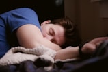 A person is sleeping peacefully on a bed, wearing a blue shirt. Their head is resting on their arm, and they are covered with a soft, fuzzy blanket. The room is dimly lit, creating a cozy and calm atmosphere.