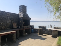 A rustic outdoor patio area with a stone grill built into a rock wall. It overlooks a calm body of water and features a couple of wooden stumps and a long wooden bench. There's a tree with branches hanging over the scene, and miscellaneous items such as a bowl and a bottle on the grill counter.