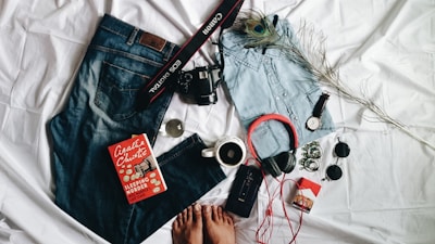 A casual arrangement of personal items displayed on a white fabric background. Includes a pair of blue jeans, a light denim shirt, a pair of headphones, a watch, a DSLR camera with a camera strap, and a book titled 'Sleeping Murder' by Agatha Christie. Accessories like sunglasses, a cup of coffee, a pack of cigarettes, and a peacock feather add to the composition. The feet of a person are partially visible at the bottom.