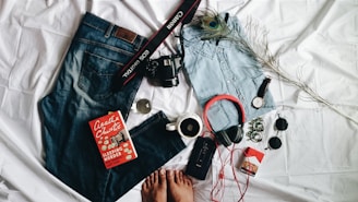 A casual arrangement of personal items displayed on a white fabric background. Includes a pair of blue jeans, a light denim shirt, a pair of headphones, a watch, a DSLR camera with a camera strap, and a book titled 'Sleeping Murder' by Agatha Christie. Accessories like sunglasses, a cup of coffee, a pack of cigarettes, and a peacock feather add to the composition. The feet of a person are partially visible at the bottom.