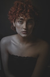 A portrait of a person with curly red hair and freckles, looking directly at the camera with a neutral expression. The background is dark, creating a dramatic contrast with the pale skin and the texture of the hair. The lighting emphasizes the facial features and shadows, adding depth to the image.