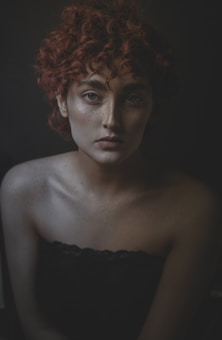 A portrait of a person with curly red hair and freckles, looking directly at the camera with a neutral expression. The background is dark, creating a dramatic contrast with the pale skin and the texture of the hair. The lighting emphasizes the facial features and shadows, adding depth to the image.