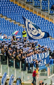 A group of enthusiastic sports fans are gathered in a stadium, holding flags, banners, and scarves that display blue and white colors and symbols. The stands are mostly empty except for this section, and the fans appear to be cheering and waving their items energetically.