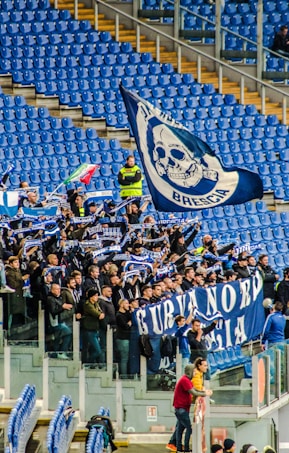 A group of enthusiastic sports fans are gathered in a stadium, holding flags, banners, and scarves that display blue and white colors and symbols. The stands are mostly empty except for this section, and the fans appear to be cheering and waving their items energetically.