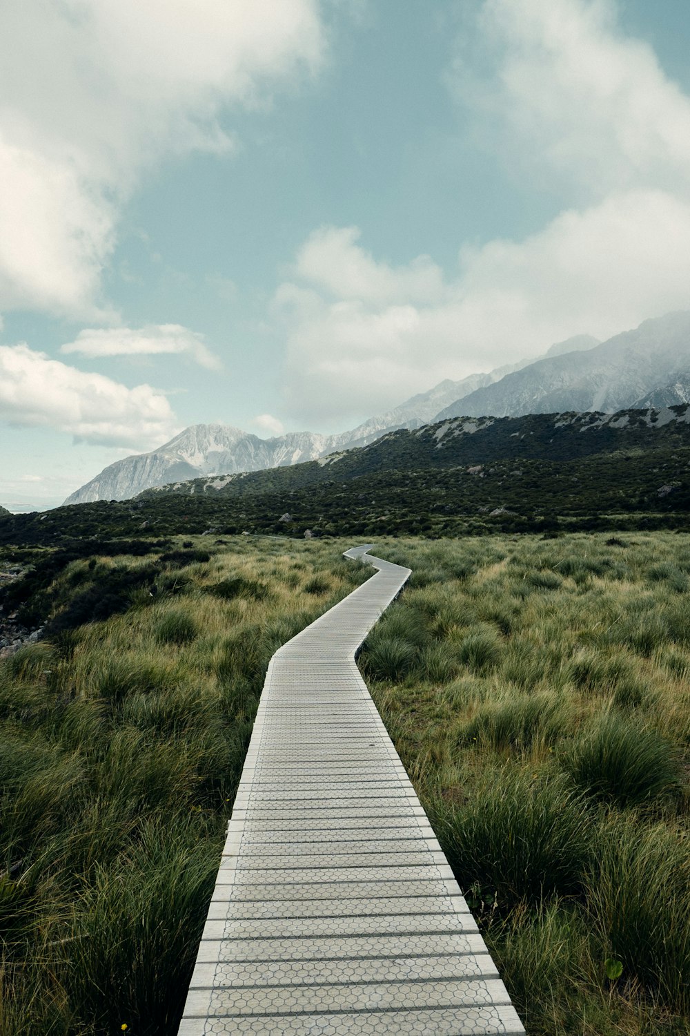 white wooden pathway on green grass field near mountains during daytime