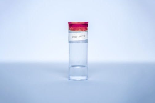 A clear glass vial with a red cap is centered in the image. A label on the vial reads '2019-NCOV'. The background is a soft gradient of blue, giving it a sterile and clinical atmosphere.