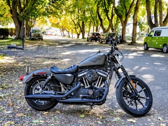 A motorcycle is parked on a tree-lined street with sunlight filtering through the leaves. The street is covered with fallen leaves and lined with parked vehicles, including a green van and several cars. The motorcycle has a matte black finish with visible branding on the fuel tank.