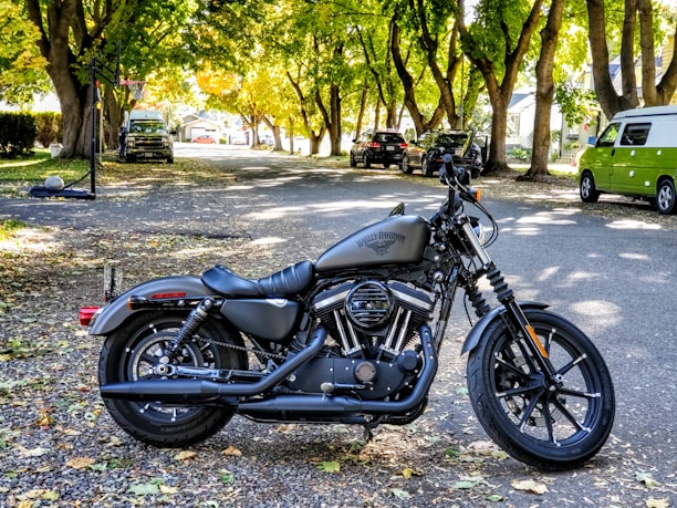 A motorcycle is parked on a tree-lined street with sunlight filtering through the leaves. The street is covered with fallen leaves and lined with parked vehicles, including a green van and several cars. The motorcycle has a matte black finish with visible branding on the fuel tank.