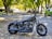 A motorcycle is parked on a tree-lined street with sunlight filtering through the leaves. The street is covered with fallen leaves and lined with parked vehicles, including a green van and several cars. The motorcycle has a matte black finish with visible branding on the fuel tank.
