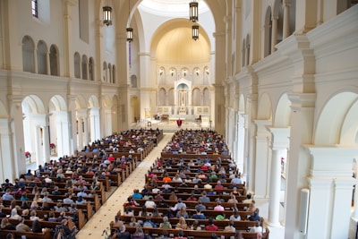 A large church interior filled with people seated in wooden pews. The architecture features high vaulted ceilings, arched windows, and detailed light fixtures. The altar area is highlighted at the front, with an ornate design and religious symbols.