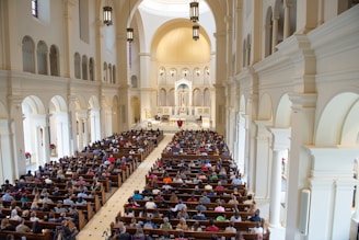 A large church interior filled with people seated in wooden pews. The architecture features high vaulted ceilings, arched windows, and detailed light fixtures. The altar area is highlighted at the front, with an ornate design and religious symbols.