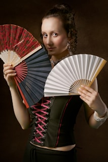A woman stands against a dark, textured background holding three different colored hand fans. She is wearing a black corset with pink laces and looks directly at the camera with a slight smile. Her curly hair is styled up, and she is also wearing a white beaded bracelet.