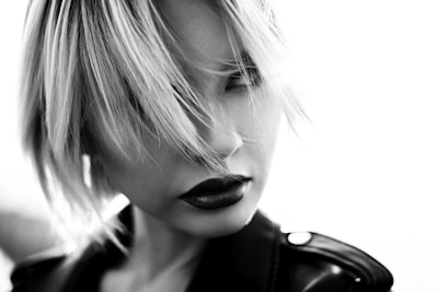 A black and white portrait of a person with short, tousled hair partially covering the face, wearing dark lipstick and a leather jacket.