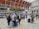 A busy train station with people gathered around a digital departure board, checking schedules. Travelers with luggage are walking through the hall, and some are sitting on benches in the background. The architectural details include a high, arched ceiling with industrial design elements.