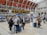 A busy train station with people gathered around a digital departure board, checking schedules. Travelers with luggage are walking through the hall, and some are sitting on benches in the background. The architectural details include a high, arched ceiling with industrial design elements.