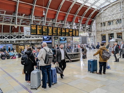 A busy train station with people gathered around a digital departure board, checking schedules. Travelers with luggage are walking through the hall, and some are sitting on benches in the background. The architectural details include a high, arched ceiling with industrial design elements.