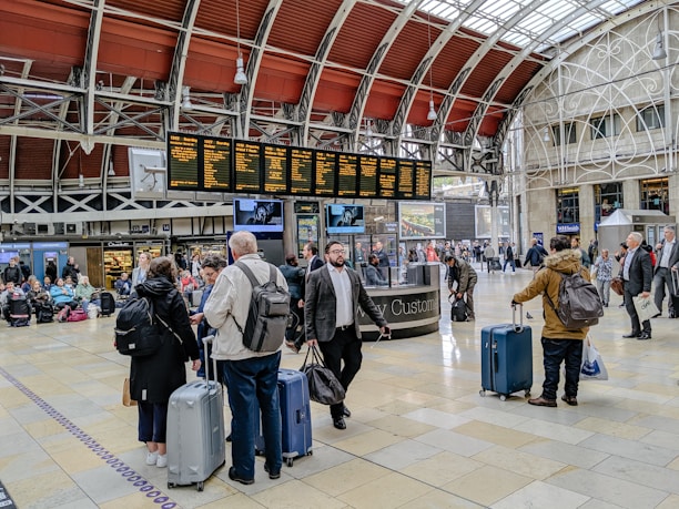 A busy train station with people gathered around a digital departure board, checking schedules. Travelers with luggage are walking through the hall, and some are sitting on benches in the background. The architectural details include a high, arched ceiling with industrial design elements.